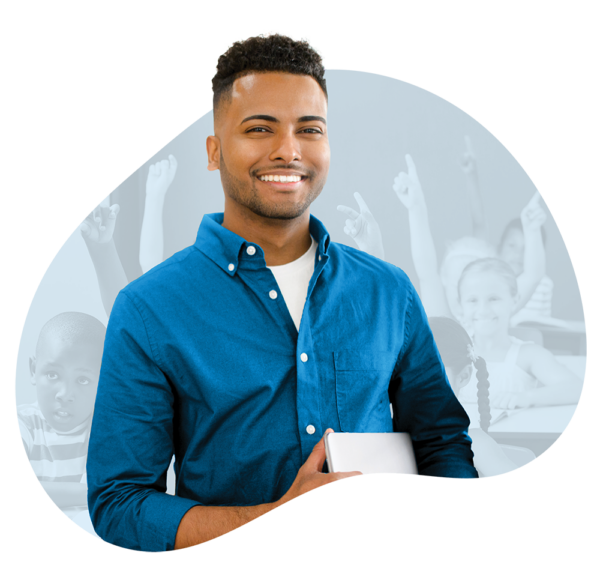 Smiling young man certified substitute teacher holding a closed laptop in front of faded background of an upper elementary school classroom